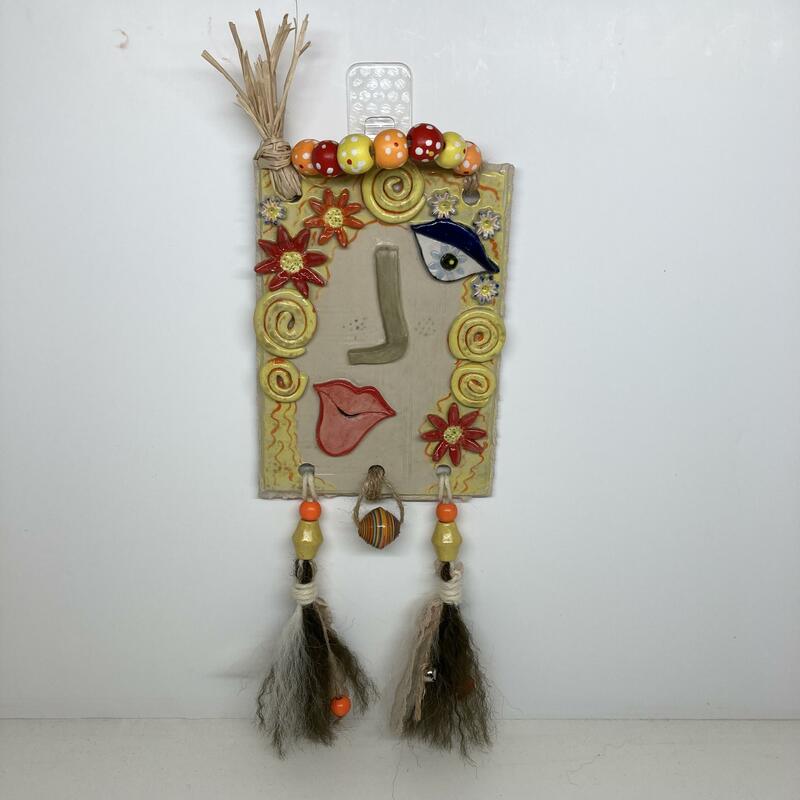 Festival. A series of ceramic and mixed media, postcard sized interpretations of well known faces. This depiction is of a summer festival goer. A stoneware tile 11x15.5cm decorated with raffia hair, wooden and paper beads and handmade tassels of lace, brushed wool and mini silver bells. In total this measures 11.5x31.5cm