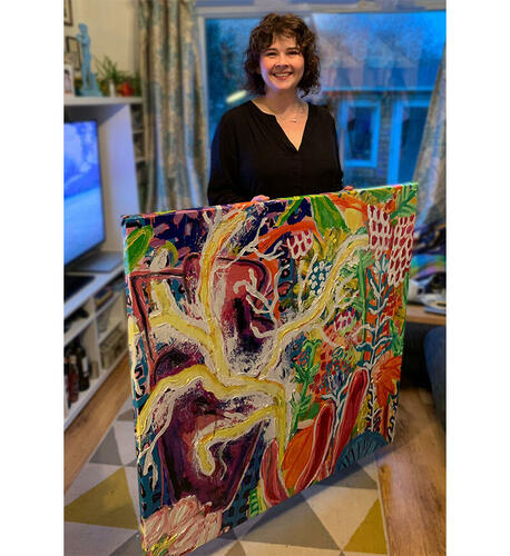 MM Plumm, a woman with curly brown hair in a black shirt standing behind a large, colourful stretched painting.