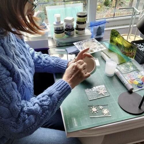 Evelyn Baxter sitting at a her workmat using glass enamels to create snowflake patterns for fused glass lanterns.