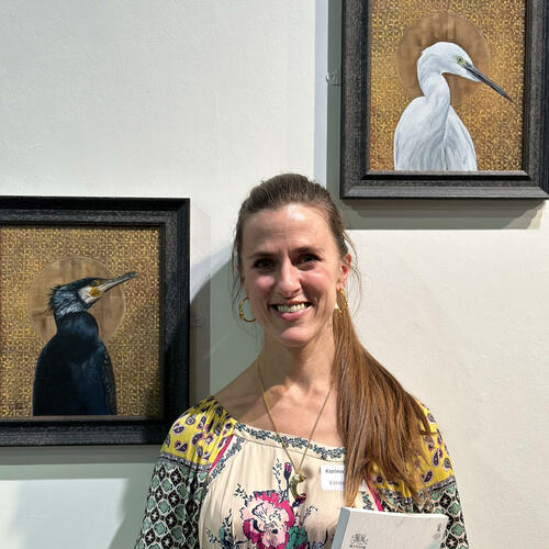 Oxford artist Karina Tarin smiling with two of her bird paintings at the Royal Society of British Artists exhibition RBA 