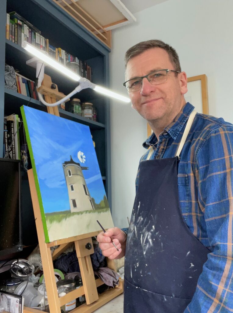 A photograph of Tim Kapp standing at his easel holding a paint brush. On the easel sits an in-progress painting of a windmill.